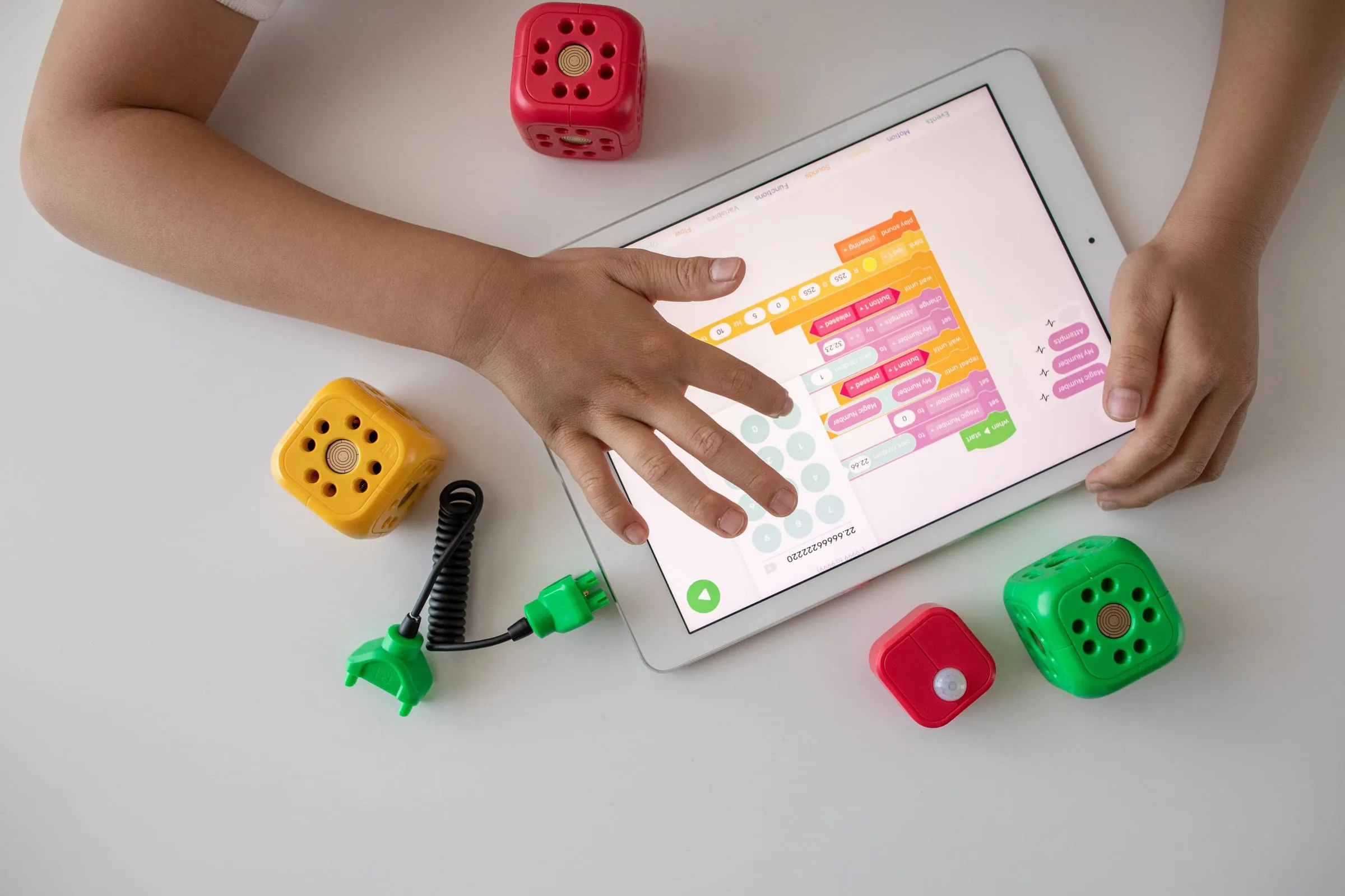 A child's hands on a tablet showing colourful code blocks, with small robot kit cubes on the table.